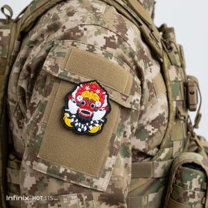 Peut inclure: Un écusson coloré représentant un masque de Barong balinais, avec des détails rouges, blancs, jaunes et noirs. L'écusson est fixé sur un uniforme militaire de camouflage, mettant en valeur un emblème culturel vibrant.