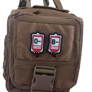 May include: Two red blood bag patches with white borders and black text. The patches are attached to a brown tactical bag. The text on the patches reads "O-" and "B-".