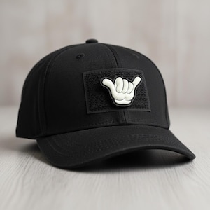 May include: Black baseball cap with a curved brim. The cap features a square patch with a white hand gesture, commonly known as the shaka sign. The cap is made of a textured fabric and has a button on top.