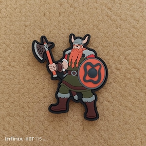 Könnte beinhalten: Eine Cartoon-Figur eines Wikingers mit rotem Bart, der eine Axt und einen Schild hält. Der Wikinger trägt einen Helm, eine grüne Tunika und braune Stiefel. Der Schild ist orange mit einem schwarzen Design. Die Figur hat einen schwarzen Umriss.