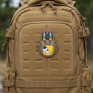 May include: A tan tactical backpack with a patch featuring a skull design wearing a helmet with night vision goggles. The patch is yellow and black, with a skull on the left side and a smiling face on the right.