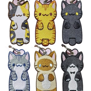 May include: Six embroidered cat keyrings with different coloured fur and patterns. Each keyring features a cute cartoon cat with a pink heart above its head and a silver key ring.