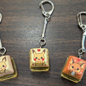 May include: Three keychains featuring Pokemon characters. Each keychain has a small, square charm with a different Pokemon character. The charms are yellow and orange with red accents. The keychains are silver.