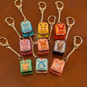 May include: A collection of keychains featuring colorful, rectangular charms with cartoon character designs. Each charm is attached to a silver-colored keyring. The charms are in various colors, including blue, yellow, orange, pink, and green.