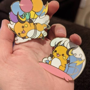 May include: Two gold-colored enamel pins featuring a cartoon yellow Pikachu holding balloons and riding a surfboard. The pins are on a hand.