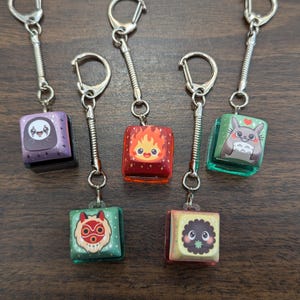 May include: Keychains featuring colorful, cube-shaped charms with cartoon character designs. Each charm is attached to a silver keyring with a spring coil. The charms have various colors, including purple, red, green, and yellow, and are displayed on a wooden surface.