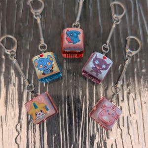May include: A selection of keyrings with cartoon character designs. Each keyring features a square, translucent charm with a different character illustration, attached to a silver keyring with a spring coil. The characters include a blue mouse, a pink shark, and a pink nurse.