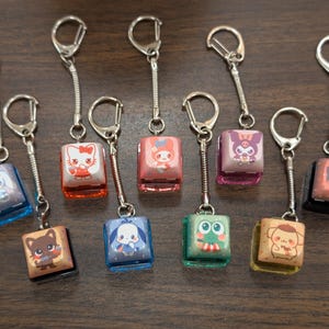 May include: Ten colorful keychains with different cartoon characters on them. Each keychain has a silver metal ring and a silver chain. The keychains are on a brown wooden surface.