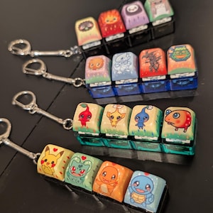 May include: Keychains featuring colorful, custom-designed keycaps with various cartoon character illustrations. Each keycap is mounted on a black base and attached to a silver keychain. The keycaps showcase a variety of characters in vibrant colors.