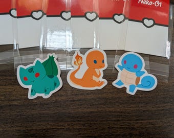 May include: Three Pokemon character stickers: a green Bulbasaur, an orange Charmander, and a blue Squirtle. The stickers are on a brown wooden surface.