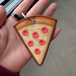 May include: A pizza slice keychain with a metal ring. The keychain is a fabric patch with a pizza slice design, featuring a crust, cheese, and six pepperoni slices. The patch is outlined with a brown border.