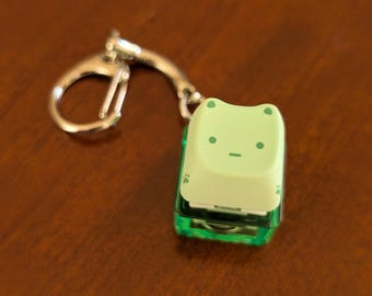 以下が含まれることがあります： シルバーのキーリングが付いた、薄緑色のカエル型キーホルダー。 キーホルダーは、黒い目と口が付いた薄緑色の顔をしています。 ボディは透明な緑色です。