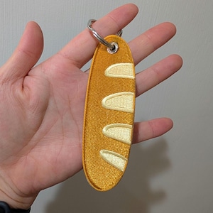 May include: An embroidered baguette-shaped keyring. The keyring is yellow with white detailing and a silver metal ring. The design resembles a loaf of bread.