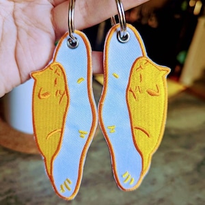 May include: Two white keychains with yellow embroidered cat designs. The cats are curled up in a sleeping position. The keychains are attached to silver key rings.