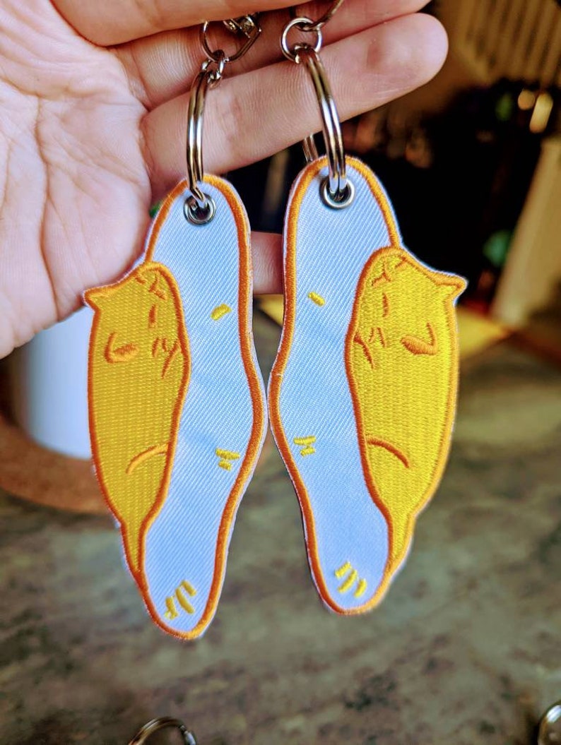 May include: Two white keychains with yellow embroidered cat designs. The cats are curled up in a sleeping position. The keychains are attached to silver key rings.