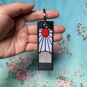 May include: Embroidered keychain with a red sun and white rays on a black background. A grey arch is at the base. A silver key ring is attached. The keychain is held in a hand.