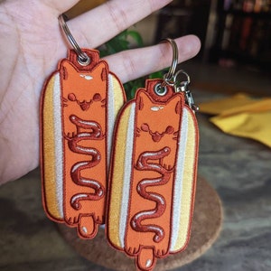May include: Two embroidered keychains shaped like hot dogs with cats inside. The cats are orange and the hot dogs are yellow with brown mustard.
