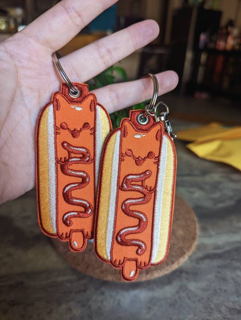 May include: Two embroidered keychains shaped like hot dogs with cats inside. The cats are orange and the hot dogs are yellow with brown mustard.