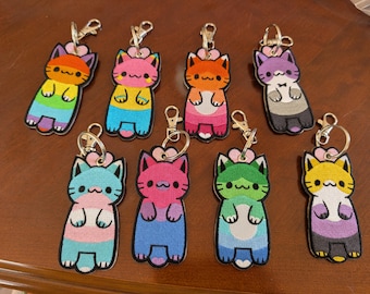 May include: A selection of embroidered cat keyrings, each featuring a different pride flag design. The keyrings depict cartoon cats with heart-shaped flowers and metal clasps. The cats are in various colours, including rainbow, pink, orange, and purple.