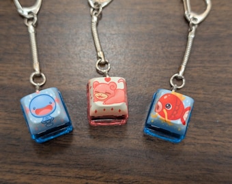 May include: Three keychains with blue and white keycaps featuring cartoon animal designs. The keycaps are attached to silver chains with a silver loop at the top. The keycaps feature a blue fish, a pink pig, and a red fish.