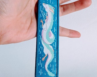 Pode incluir: Um chaveiro de tecido azul com um design de dragão branco bordado. O dragão está enrolado e tem um esquema de cores azul claro e rosa.