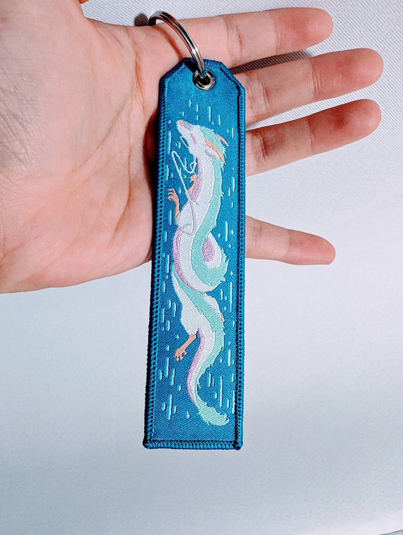 May include: A blue fabric keychain with a white embroidered dragon design. The dragon is curled up and has a light blue and pink color scheme.