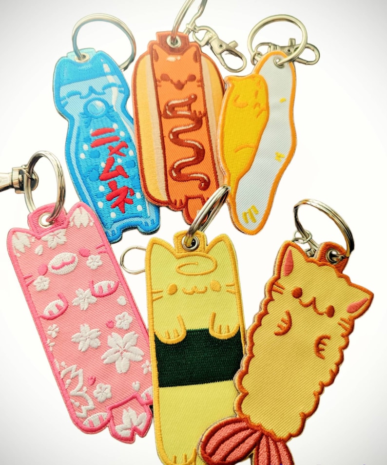 May include: A set of six colorful embroidered keychains featuring a variety of Japanese-inspired designs, including a hot dog, a bottle of soda, a sushi roll, and three cats. The keychains are made of fabric and have a cute and whimsical style.
