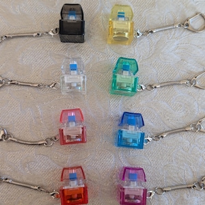 May include: A selection of small, colourful keychains. Each keychain has a small, rectangular, transparent case with a coloured top and a silver chain. The colours include black, yellow, clear, green, red, blue, orange, and pink.