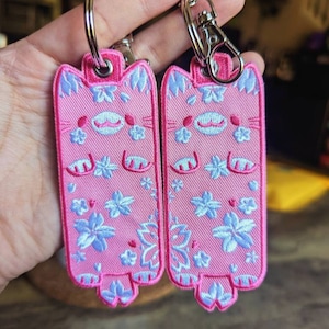 May include: Two pink cat-shaped keychains with blue floral embroidery and silver key rings. The keychains have a cute, cartoon-like design.