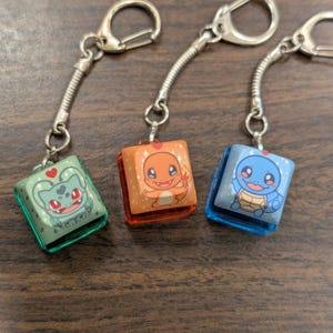 May include: Three keychains featuring Pokemon characters. The keychains are made of clear plastic with a square shape. The first keychain features a green Bulbasaur, the second keychain features an orange Charmander, and the third keychain features a blue Squirtle.