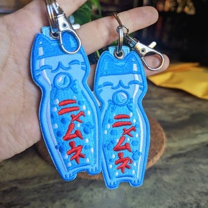 May include: Two blue and white embroidered cat bottle keychain charms with red Japanese characters. The charms are attached to silver key rings.