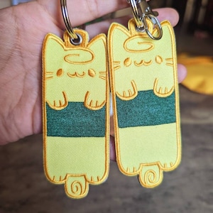 May include: Two yellow cat-shaped keychains with green accents and orange eyes. The keychains are attached to silver key rings.