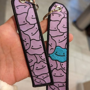 May include: Two purple keychains with black outlines and a pattern of smiling faces. One keychain has a single blue smiling face in the pattern.