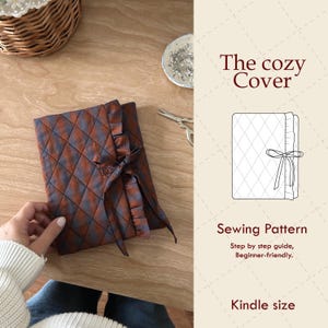 Könnte beinhalten: Gesteppte Kindle-Hülle in Braun- und Blautönen mit Rautenmuster und dekorativer Schleife. Die Hülle hat einen gerüschten Rand und wird auf einer Holzoberfläche gezeigt. Text: "The cozy Cover" und "Sewing Pattern".
