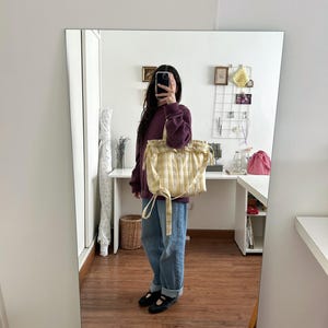 May include: A yellow and white checkered tote bag with a drawstring closure. The bag is held by a person wearing a burgundy sweatshirt and blue jeans. The bag has a soft, quilted appearance.