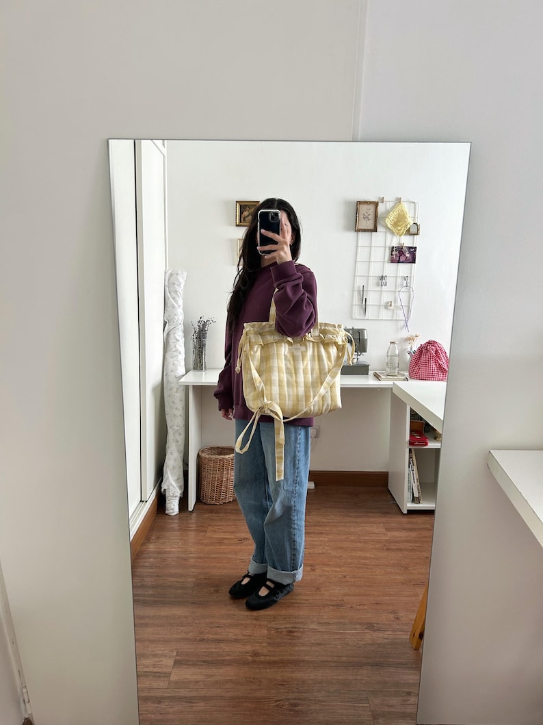 May include: A yellow and white checkered tote bag with a drawstring closure. The bag is held by a person wearing a burgundy sweatshirt and blue jeans. The bag has a soft, quilted appearance.