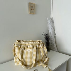 May include: A yellow and white gingham tote bag with ruffled trim and long handles. The bag has a small rectangular label and is sitting on a white surface. A pair of scissors rests in front of the bag.