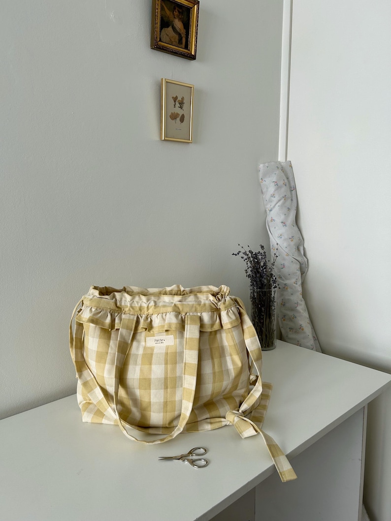 May include: A yellow and white gingham tote bag with ruffled trim and long handles. The bag has a small rectangular label and is sitting on a white surface. A pair of scissors rests in front of the bag.