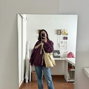 May include: A person wearing a dark purple sweatshirt, light blue jeans, and black shoes. A yellow and white checkered drawstring bag is worn over the shoulder. The person is holding a phone in front of their face, taking a mirror selfie.
