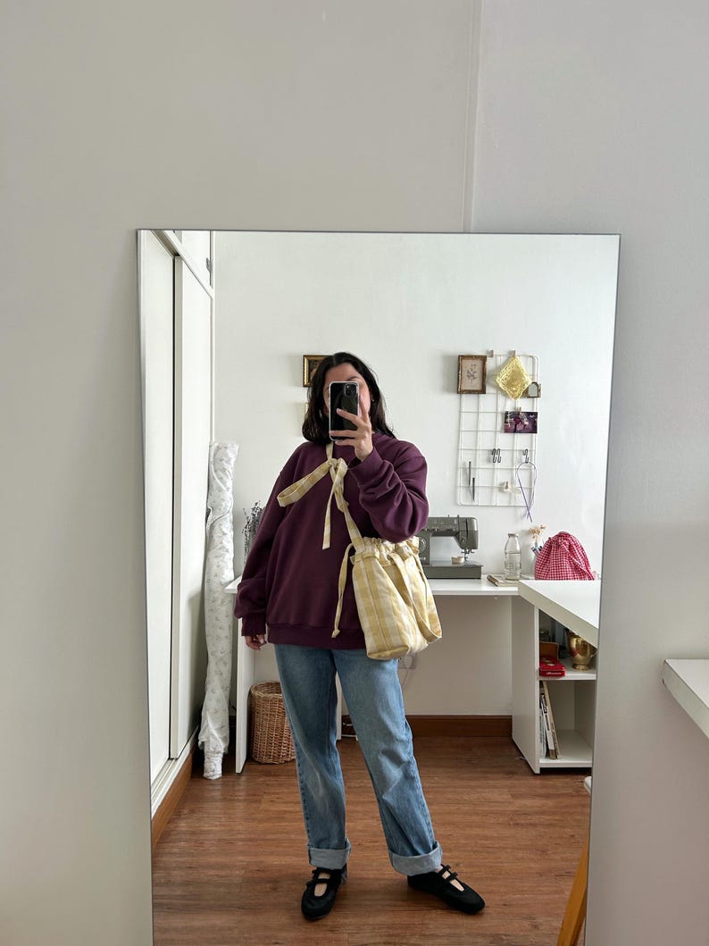 May include: A person wearing a dark purple sweatshirt, light blue jeans, and black shoes. A yellow and white checkered drawstring bag is worn over the shoulder. The person is holding a phone in front of their face, taking a mirror selfie.