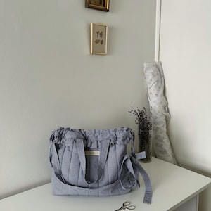 May include: A blue and white gingham tote bag with a gathered top and shoulder strap sits on a white surface. A small pair of scissors and a vase of lavender are nearby. Two framed pictures hang on the wall behind the bag.