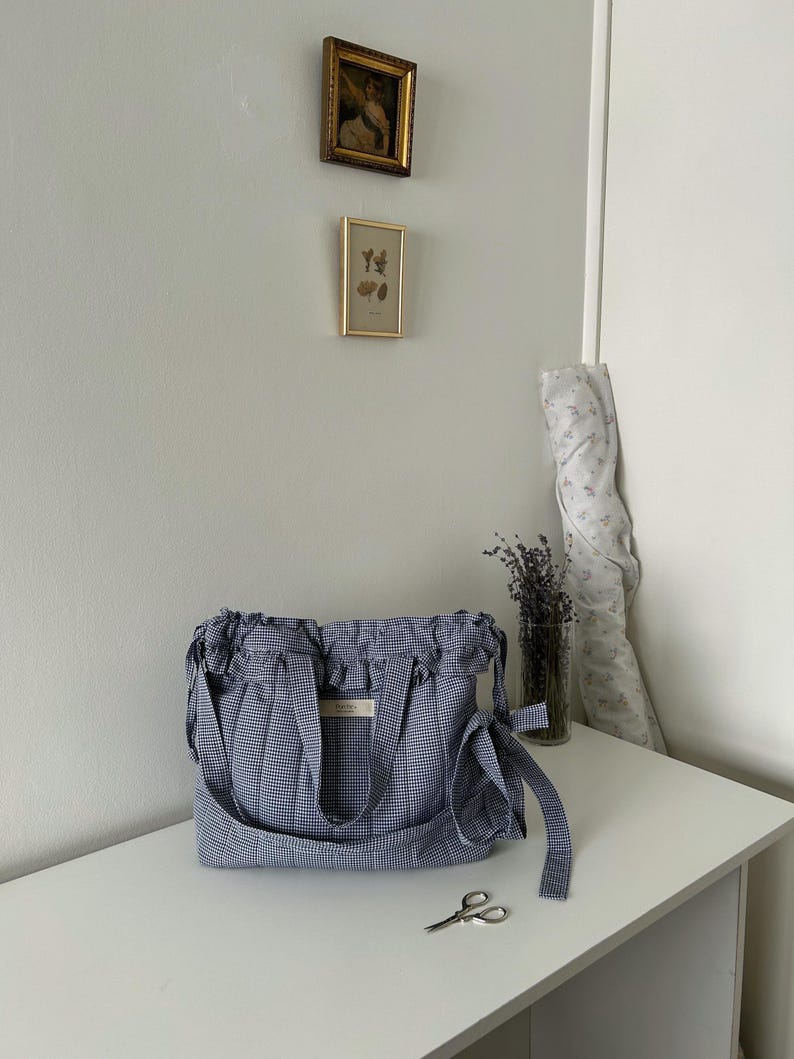 May include: A blue and white gingham tote bag with a gathered top and shoulder strap sits on a white surface. A small pair of scissors and a vase of lavender are nearby. Two framed pictures hang on the wall behind the bag.