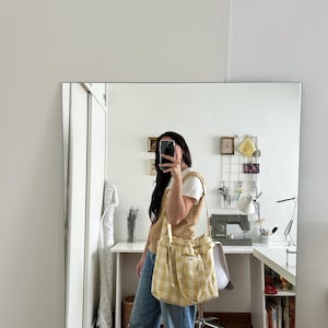 May include: A yellow and white plaid tote bag with a drawstring closure and a shoulder strap. The bag is held by a person wearing a tan knit vest, a white t-shirt, and blue jeans. The background includes a mirror and a desk.