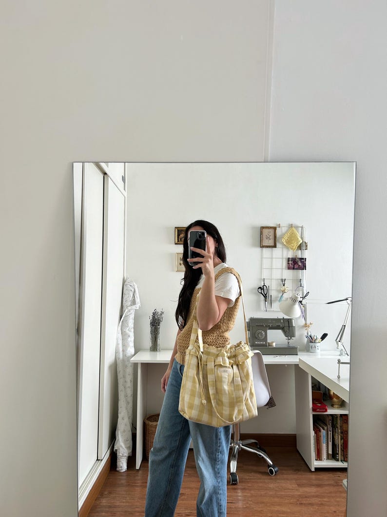 May include: A yellow and white plaid tote bag with a drawstring closure and a shoulder strap. The bag is held by a person wearing a tan knit vest, a white t-shirt, and blue jeans. The background includes a mirror and a desk.
