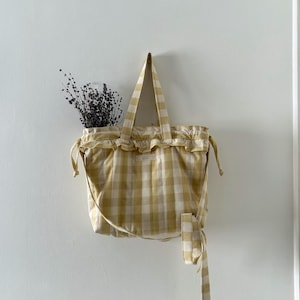 May include: A yellow and white gingham tote bag with a ruffled top and long handles hangs on a white wall. The bag has a drawstring closure and a long tie. Dried flowers are visible inside the bag.