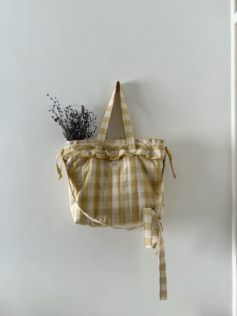 May include: A yellow and white gingham tote bag with a ruffled top and long handles hangs on a white wall. The bag has a drawstring closure and a long tie. Dried flowers are visible inside the bag.