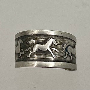 May include: Silver ring with a design of three horses running in a circle. The ring is made of a textured metal and has a brushed finish.