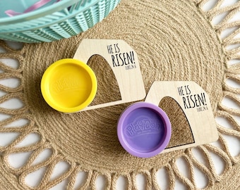 May include: Two wooden Easter egg holders with the text "He is Risen! Luke 24:6" on each. The holders are on a woven mat and each has a Play-Doh container inside. One container is yellow and the other is purple.