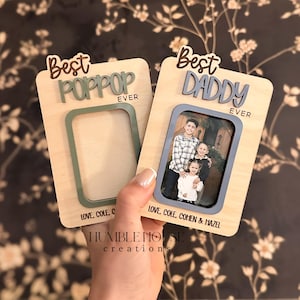 May include: Two wooden photo frames, one with the text "Best POPPOP EVER" and the other with "Best DADDY EVER." The "Best DADDY" frame holds a family photo. Both frames have a rectangular photo opening with a colored border. The frames are held in front of a floral wallpaper.
