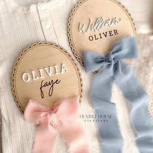 May include: Two wooden name plaques with scalloped edges, one with "Olivia Faye" and a pink ribbon, the other with "William Oliver" and a blue ribbon. The plaques are on a white onesie. The text "HUMBLE HOUSE CREATIONS" is visible.